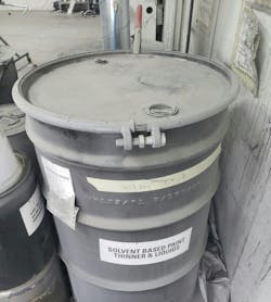 This drum of liquid solvent waste is properly labeled and grounded, as shown by the red clamp at the rear.